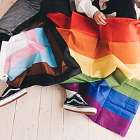 Zu sehen ist eine Regenbogenflagge die &uuml;ber den Beinen von zwei Menschen ausgebreitet liegt.