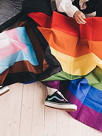 Zu sehen ist eine Regenbogenflagge die &uuml;ber den Beinen von zwei Menschen ausgebreitet liegt.