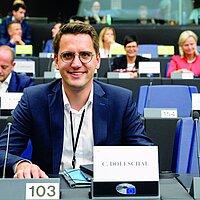 Christian Doleschal sitzt im EU-Parlament an seinem Platz, vor ihm ein Mikrofon und sein Namensschild. Er l&auml;chelt in die Kamera.