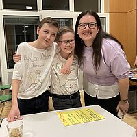 Zwei Sch&uuml;ler tragen ein T-Shirt, auf das "Anton-Miller-Restaurant" geschrieben ist und stehen mit der Betreuerin am Tisch.