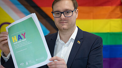 Eine Person vor einer Regenbogenflagge, in seinen H&auml;nden das Titelblatt der ver&ouml;ffentlichten Studie