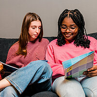 Zwei M&auml;dchen sitzen auf der Couch und schauen sich U18-Infomaterial an. 