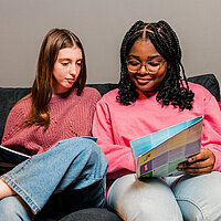 Zwei M&auml;dchen sitzen auf der Couch und schauen sich U18-Infomaterial an. 