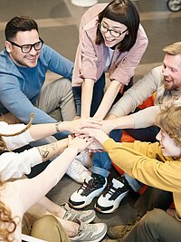 Eine Gruppe von Mitarbeitenden sitzt auf dem Boden im Kreis. Alle halten ihre H&auml;nde zu einer motivierenden Geste in die Mitte.