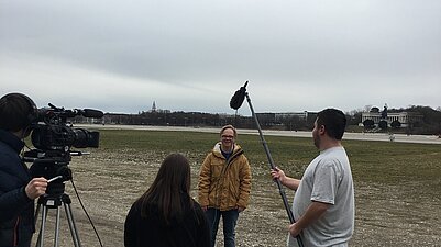 Ein junger Mann steht vor einer Kamera in einem gelebn Anorak. Ein Jugendlicher h&auml;lt das Mikrofon.