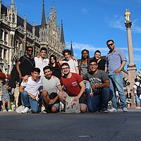 11 internationale Jugendliche stehen zum Gruppenfoto zusammen auf dem M&uuml;nchner Marienplatz. Sie haben sich gegenseitig die Arme &uuml;ber die Schultern gelegt und l&auml;cheln in die Kamera.
