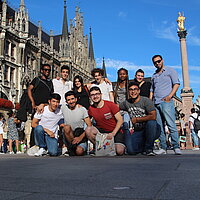 11 internationale Jugendliche stehen zum Gruppenfoto zusammen auf dem M&uuml;nchner Marienplatz. Sie haben sich gegenseitig die Arme &uuml;ber die Schultern gelegt und l&auml;cheln in die Kamera.