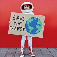 Ein M&auml;dchen in einem Astronautenkost&uuml;m mit Helm steht vor rotem Hintergrund und h&auml;lt ein selbsgebasteltes Schild in den H&auml;nden mit einer Weltkugel und dem Text "Save the Planet"