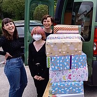 Vor einem Kastenwagen t&uuml;rmen sich eingepackte Geschenke, zwei Frauen stehen l&auml;chelnd dabei.