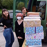 Vor einem Kastenwagen t&uuml;rmen sich eingepackte Geschenke, zwei Frauen stehen l&auml;chelnd dabei.