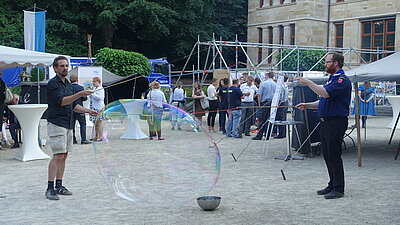 Im Vordergrund machen zwei M&auml;nner riesige Seifeblasen. Im Hintergrund unterhalten sich Menschen.