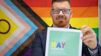 Eine Person h&auml;lt die Studie in die Kamera, im Hintergrund eine Regenbogenflagge