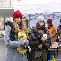Zwei junge warm angezogene Frauen haben Wolldecken in der Hand, dahinter der Ausgabe-Tisch f&uuml;r die Wolldecken.