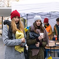 Zwei junge warm angezogene Frauen haben Wolldecken in der Hand, dahinter der Ausgabe-Tisch f&uuml;r die Wolldecken.
