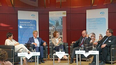 5 Podiumsteilnehmende sitzen im Halbkreis auf dem Podium