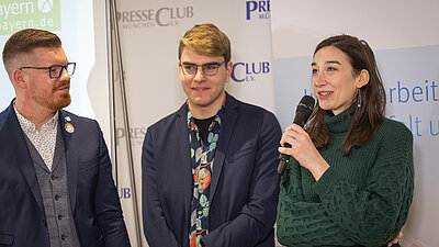 Drei Personen auf dem Bild, eine Person rechts auf dem Bild spricht in ein Mikrofon