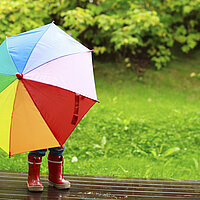 Ein bunt angezogenes Kind in Regenstiefeln und Regenkleidung, Gesicht und Oberk&ouml;rper sind durch einen bunten Regenschirm verdeckt, der es vor dem Regenbsch&uuml;tzt. 