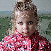Ein blondes M&auml;dchen in einer rot-gebl&uuml;mten Jacke schaut traurig in die Kamera.