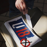 Eine Hand steckt einen Wahlzettel in eine graue Wahlunre mit einem großen Aufkleber mit dem Logo der U18-Wahl