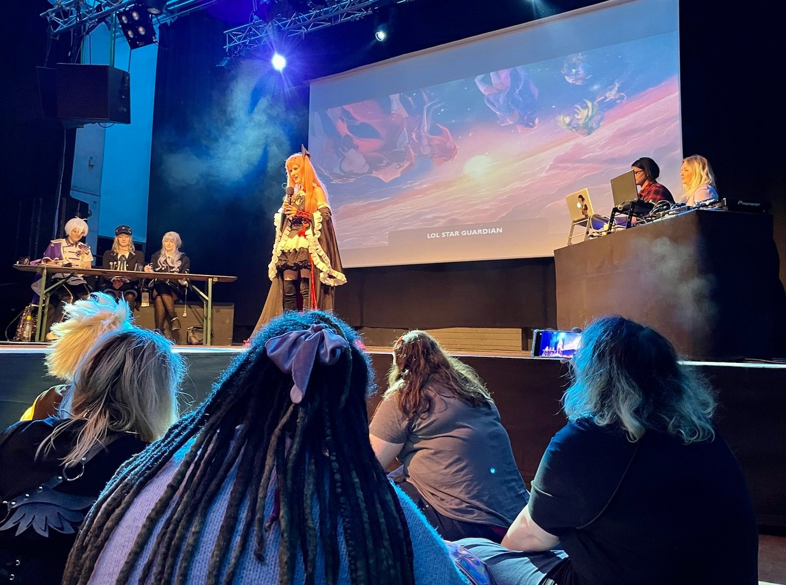  'eine Cosplayerin h&auml;lt bei derPreisverleihung des Bayerischen Gamespreises eine Rede auf einer B&uuml;hne. Junge Menschen h&ouml;ren sitzen im Publikum. Link zu www.gamesfestival.de/